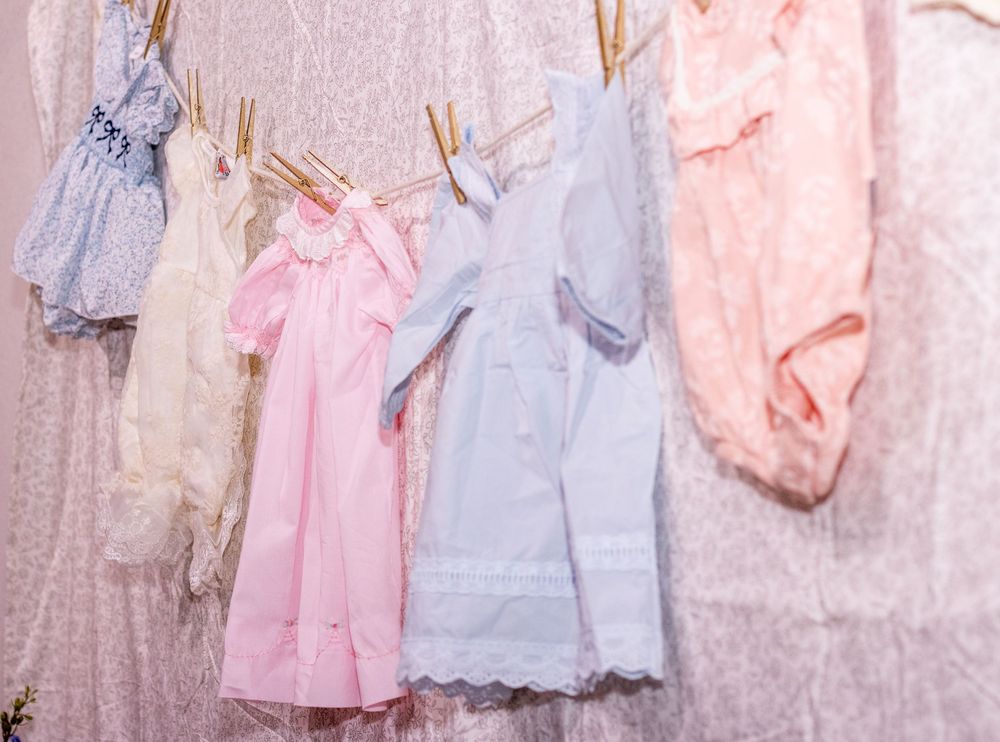 Vintage baby dresses and clothing in soft pastel colors hang on a white wall display.