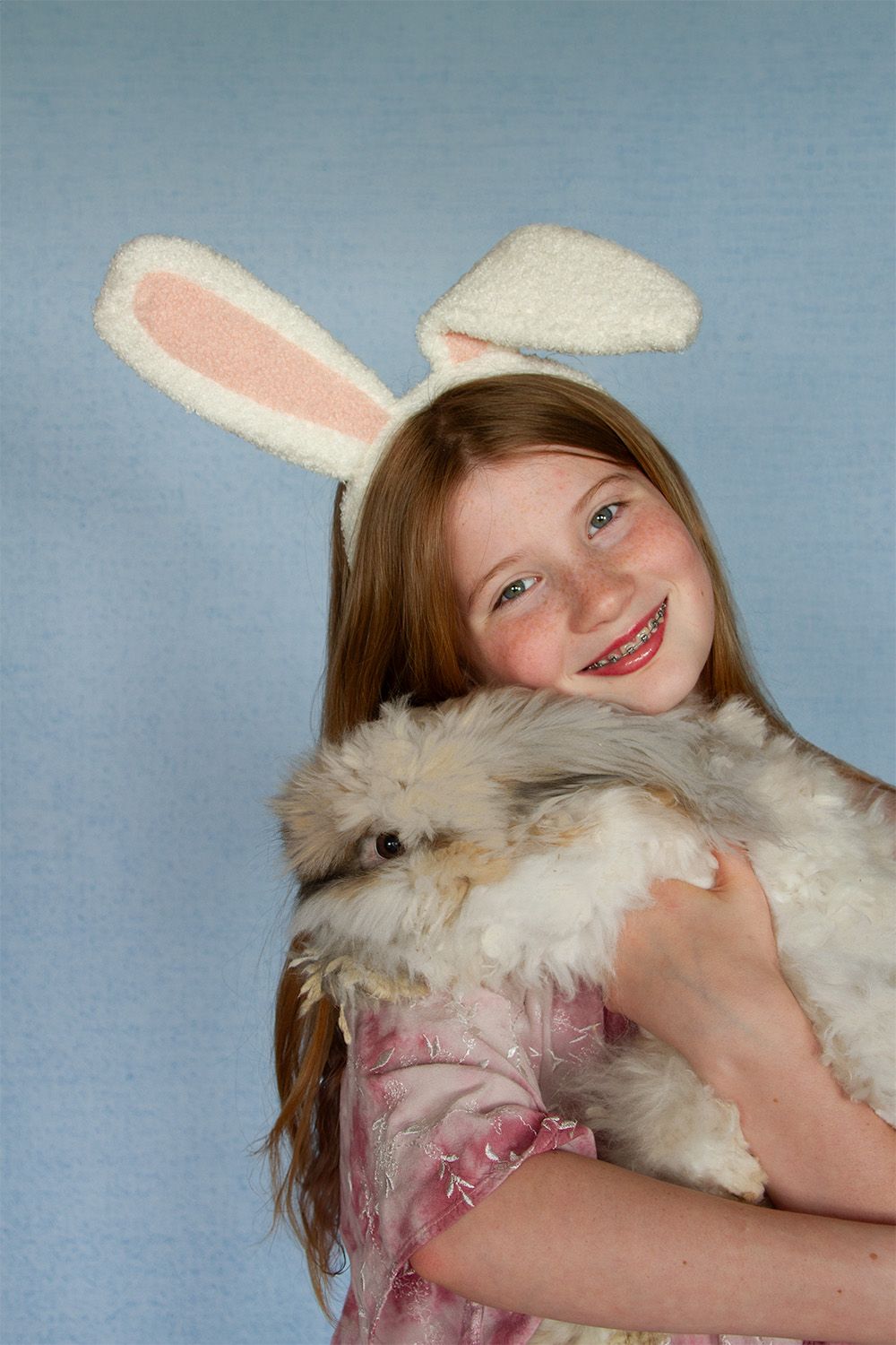 little girl wearing a rabbit ears headband and holding a fluffy gray rabbit in front of a blue backdrop- taken by Delta Blossoms Photography