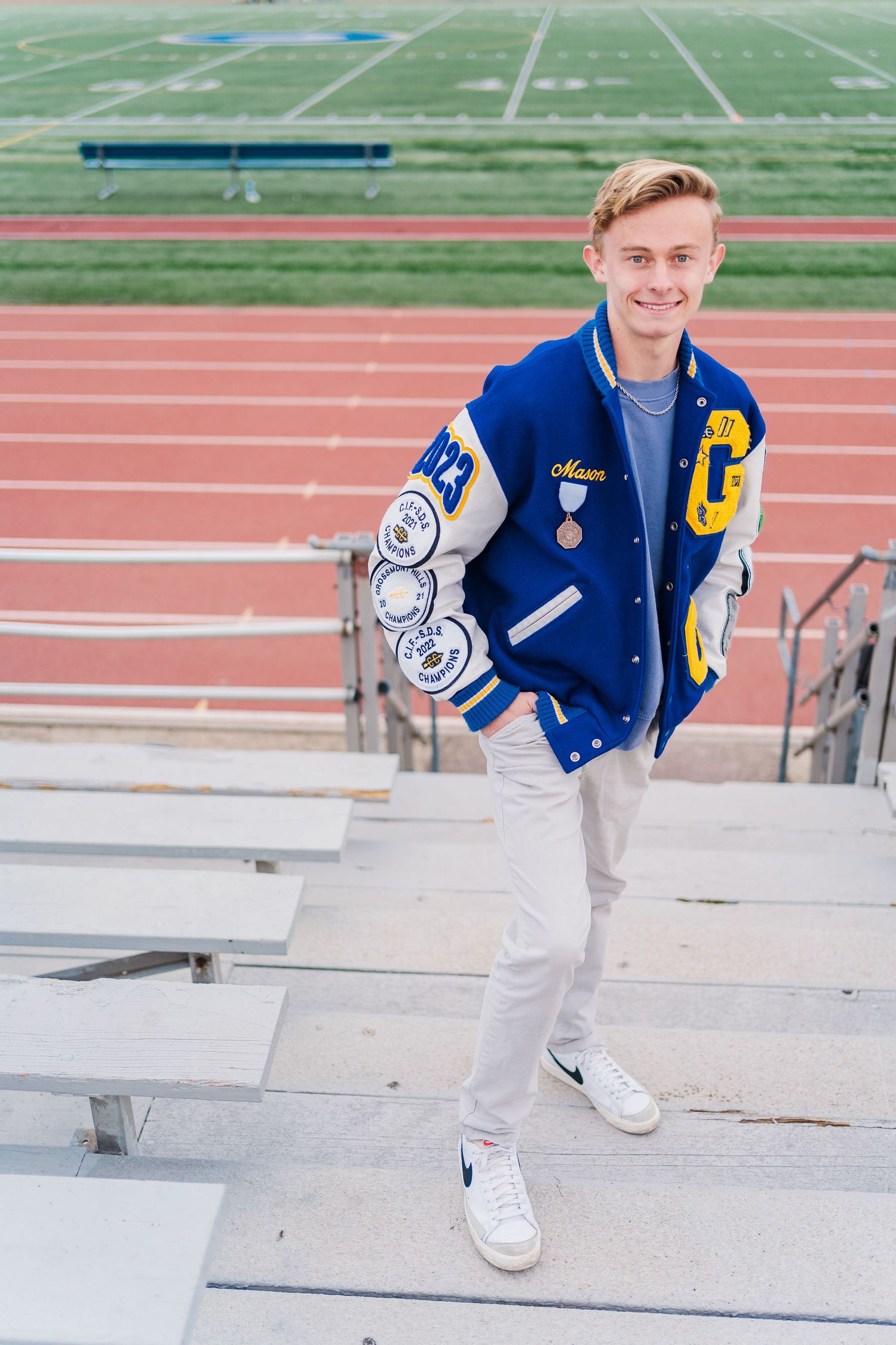 Student in blue varsity letterman jacket and white pants poses on bleachers at track and field stadium.