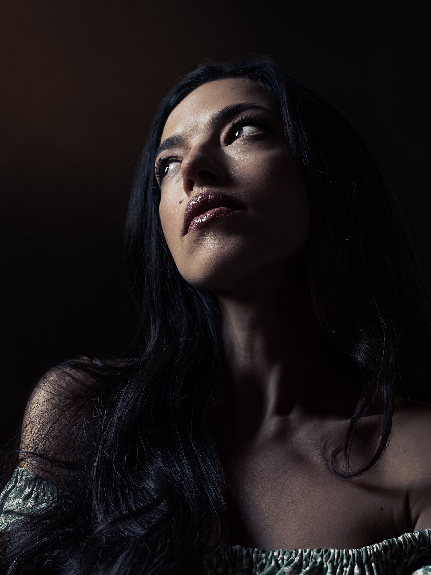 A woman with long dark hair gazes upward dramatically in moody, dark studio lighting.