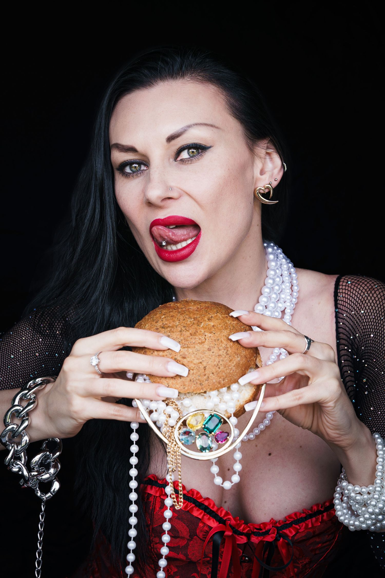 Woman in red corset and pearl necklace holding a large burger with mouth open, wearing bold red lipstick.
