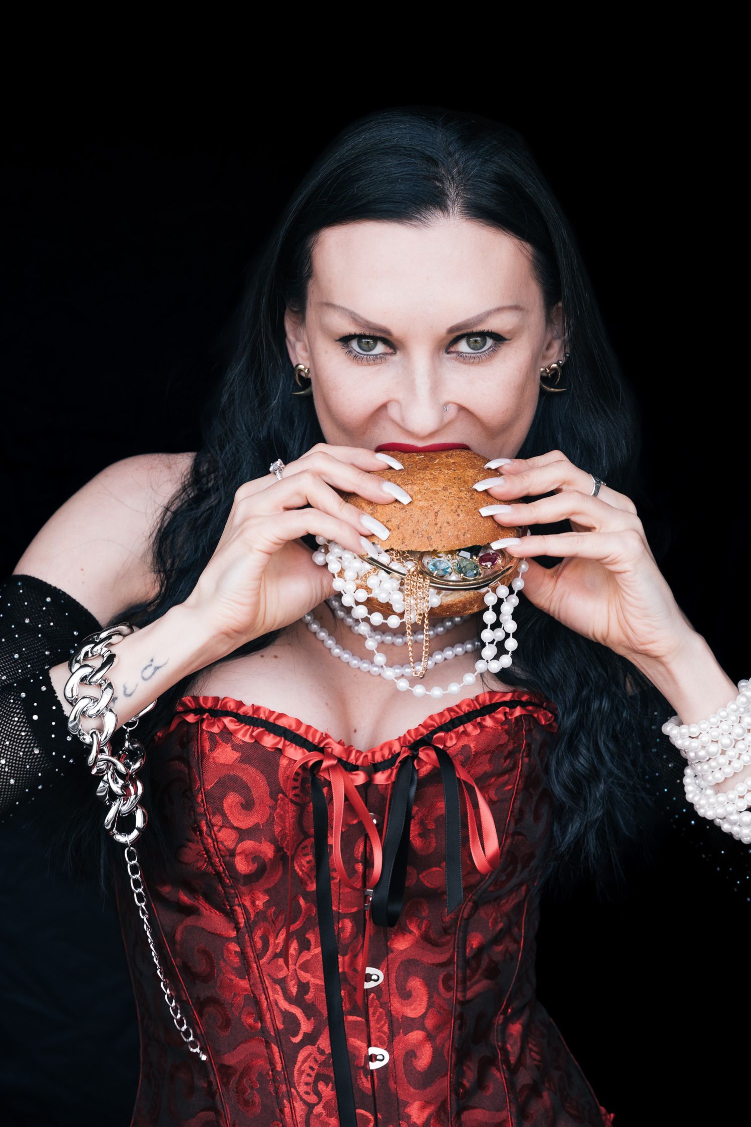 Woman in red corset and pearls eating a burger against black background.