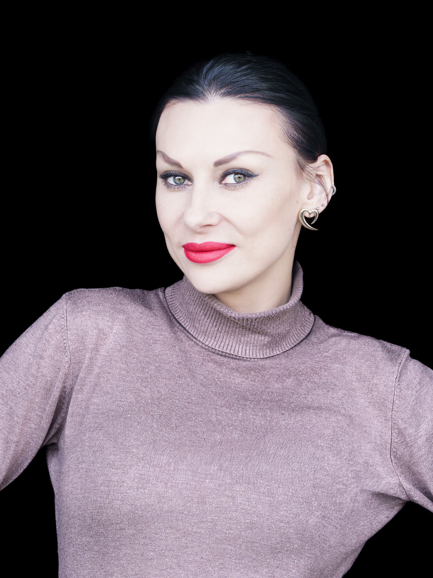 Woman with bold red lips and blue eyes wearing a mauve turtleneck against black background.