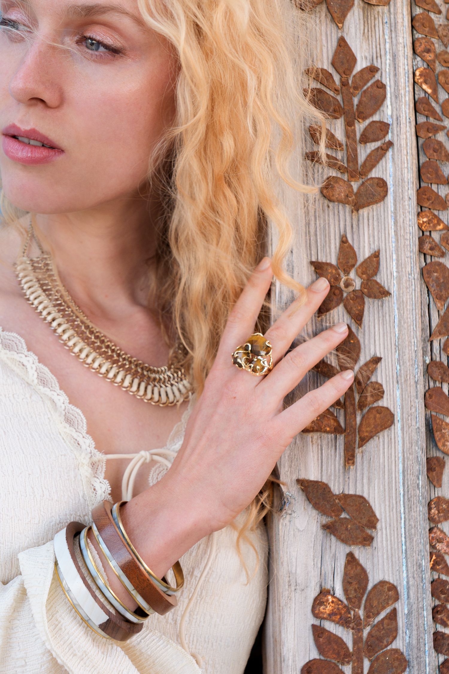Blonde woman wearing gold statement necklace, stacked bracelets, and floral ring poses against rustic wooden door.