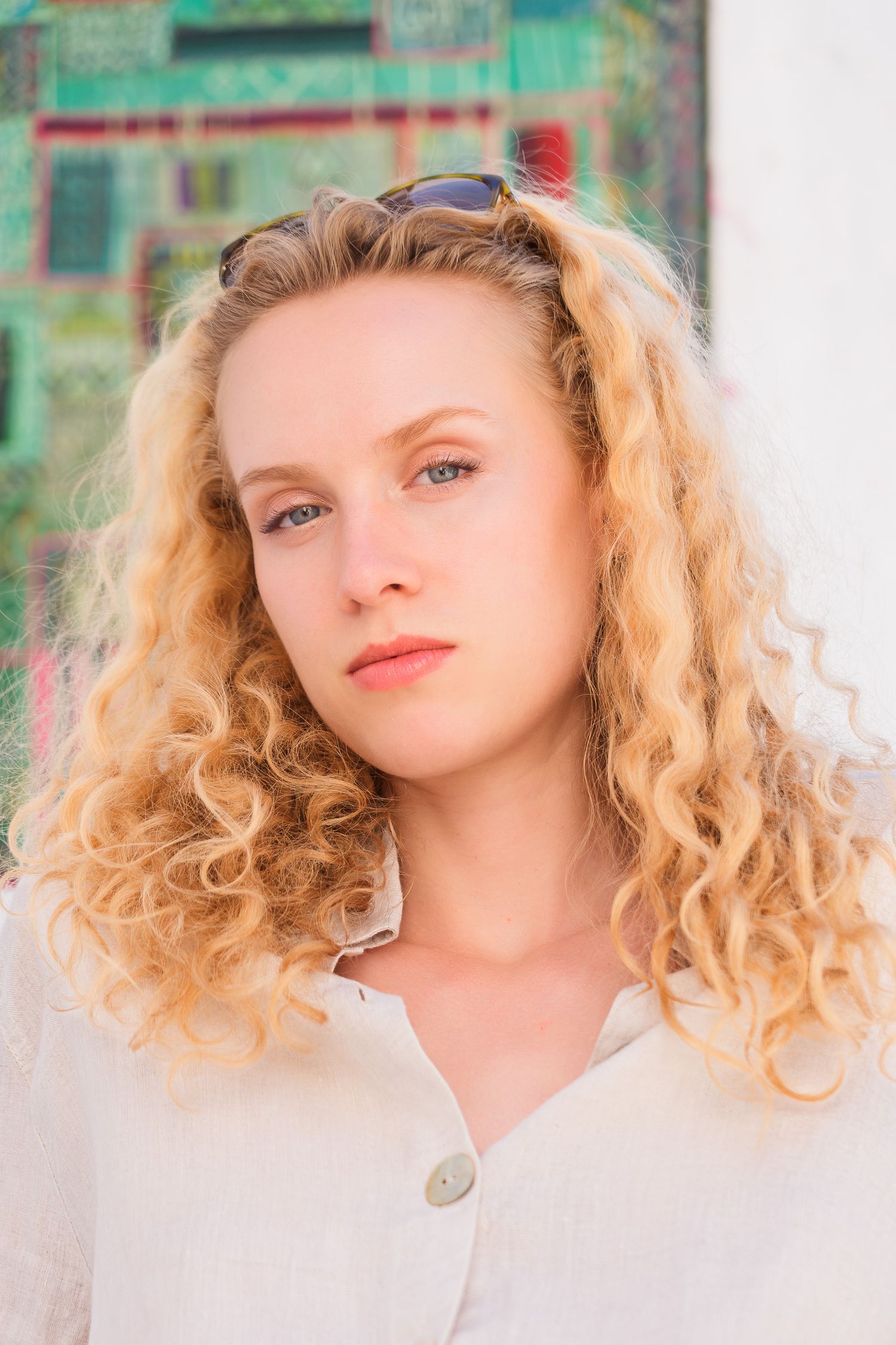 Young woman with curly blonde hair and sunglasses on head posing outdoors in white shirt.