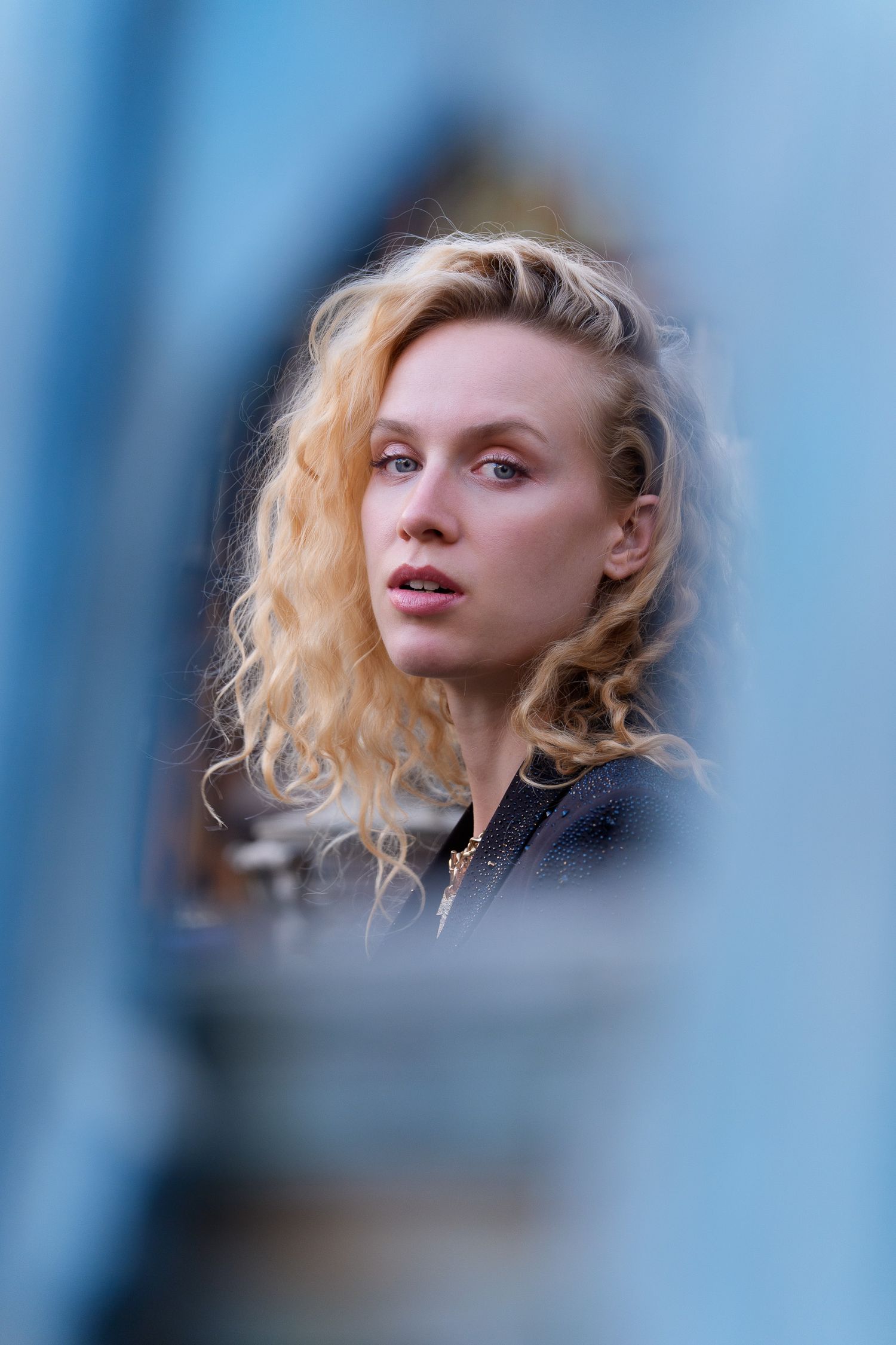 Young blonde woman with curly hair gazes thoughtfully through a blue metal frame outdoors.
