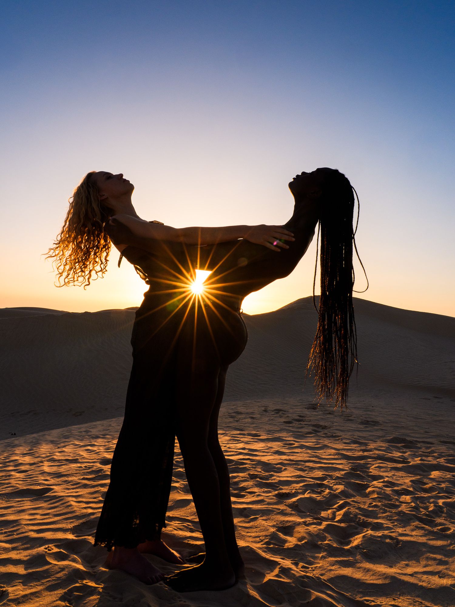 Two silhouetted dancers pose dramatically against a stunning desert sunset with golden sand dunes.