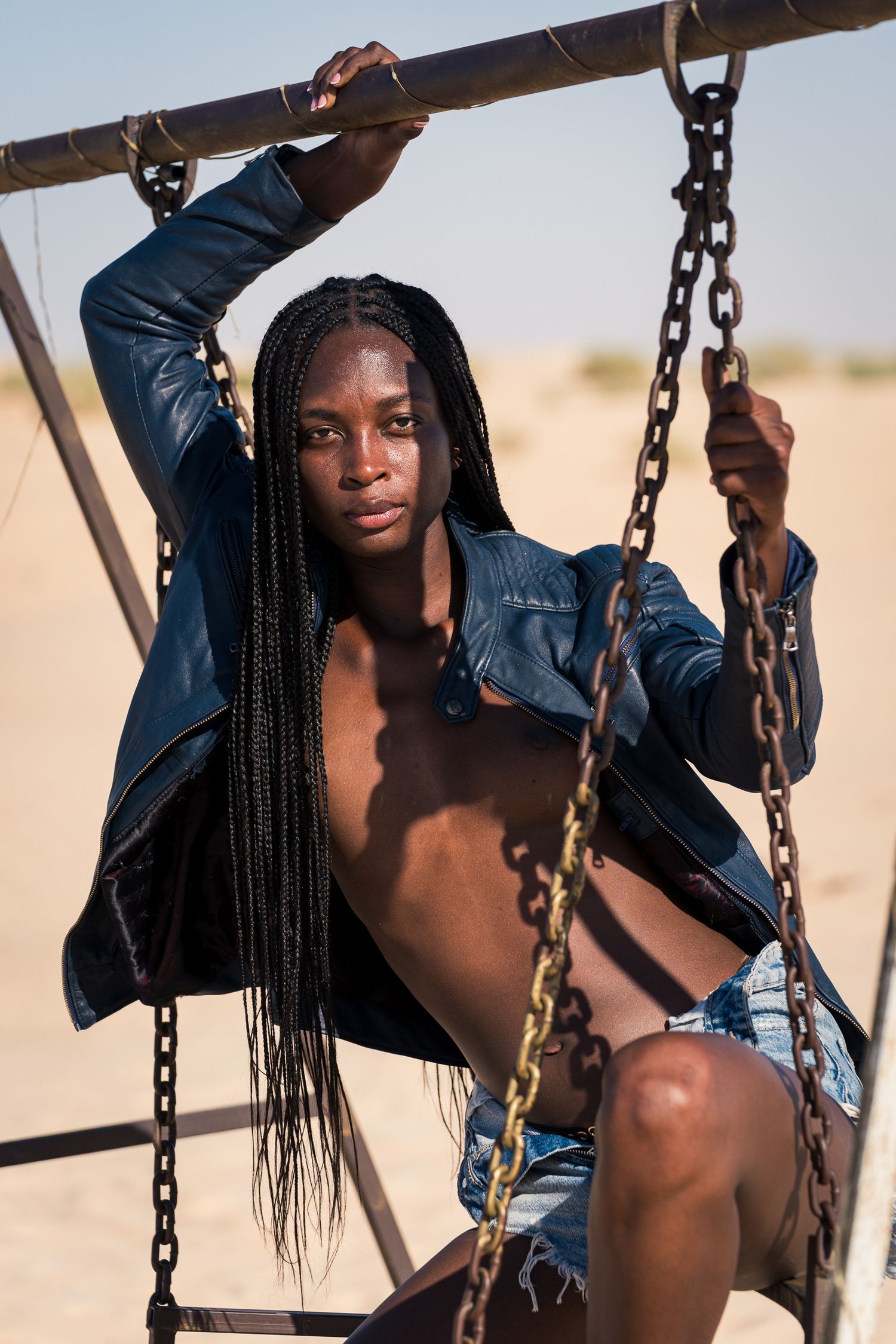 Young Black woman with long braids in leather jacket and denim shorts sitting on rustic chain swing in desert.