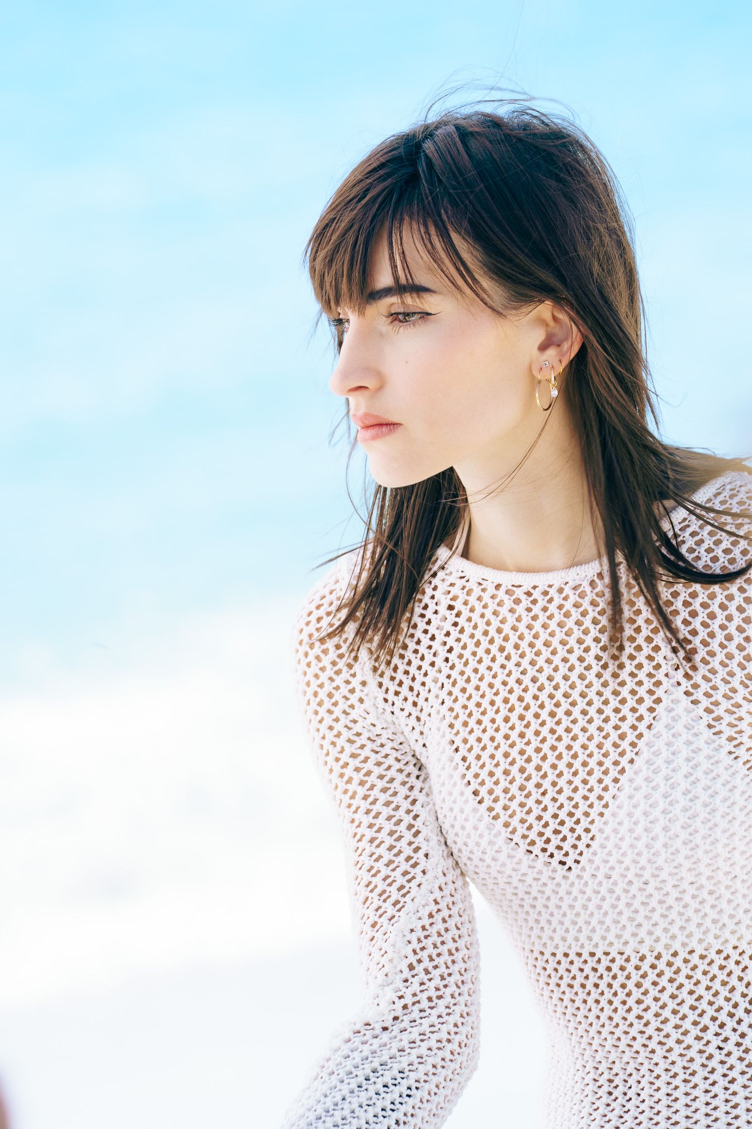 Young woman with dark hair in white mesh top gazing away at the beach on a sunny day.