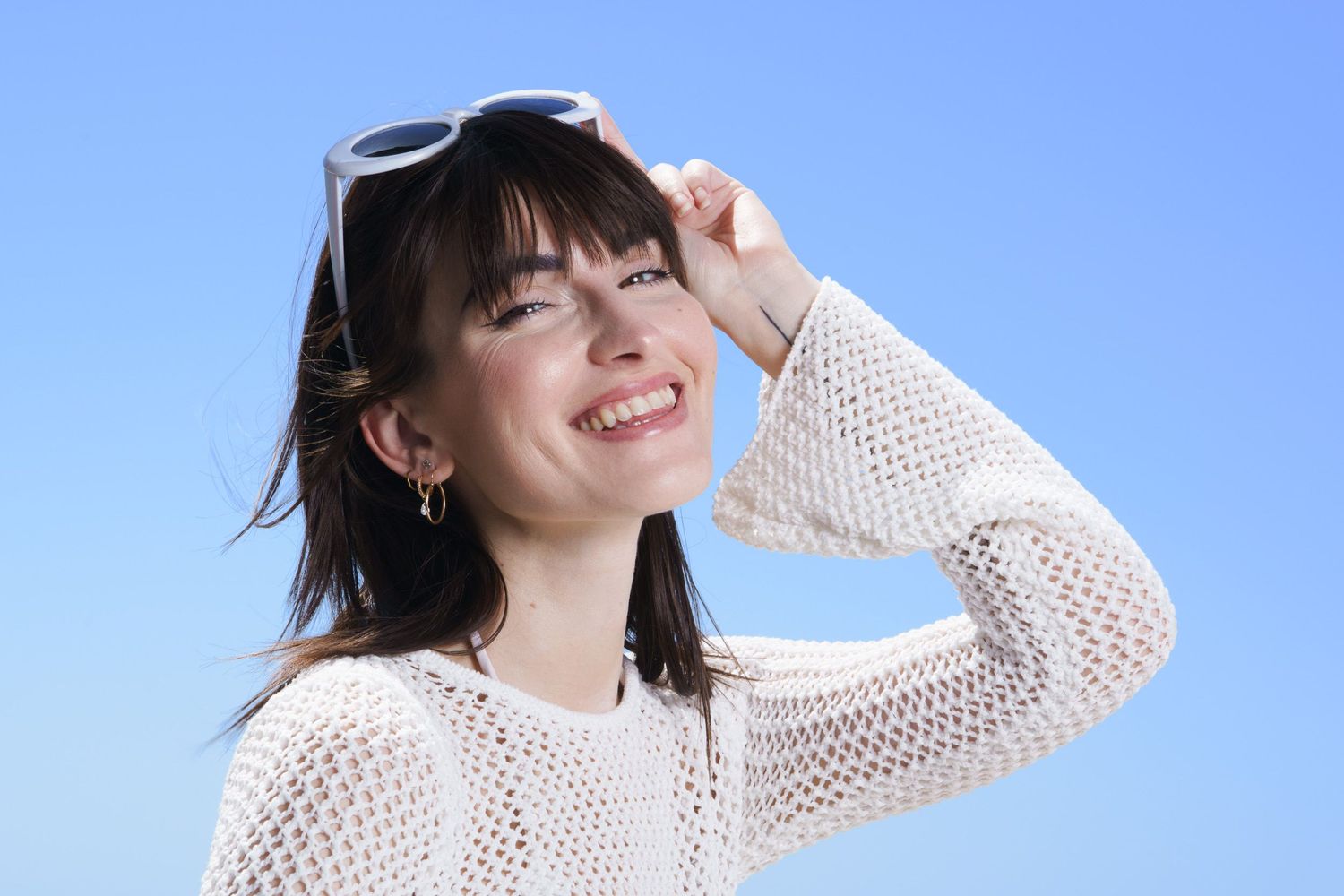 Smiling young woman with sunglasses on head, wearing white knit sweater, posing against clear blue sky.