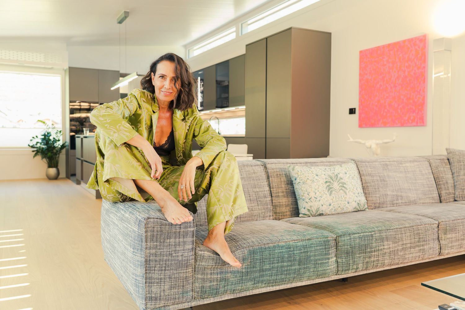 Woman in green silk outfit sitting on a modern gray sofa in a stylish open-plan living space.