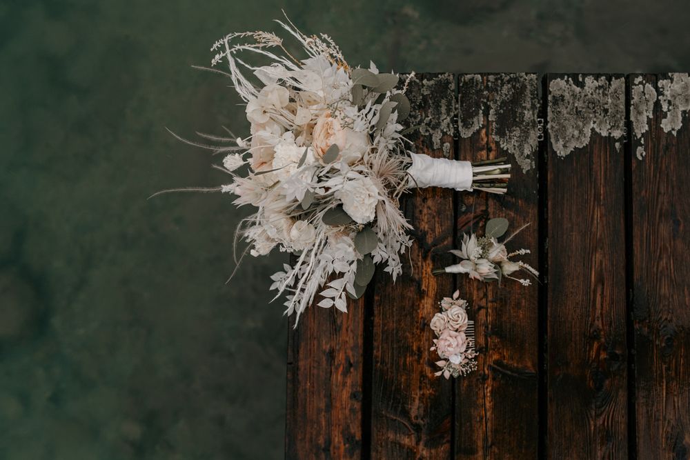 Elegant white bridal bouquet and boutonniere arranged on rustic wooden dock over green water.