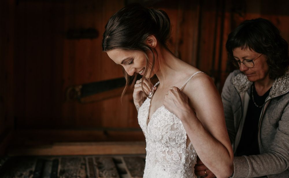 A mother helps her daughter button up a lace wedding dress in a rustic wooden interior.