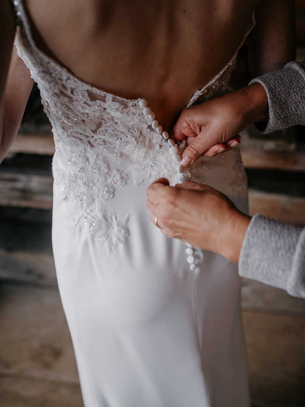 Close-up of hands fastening delicate pearl buttons on the back of a lace wedding dress.