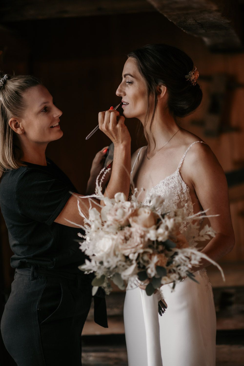 A makeup artist applies lipstick to a bride holding a bohemian white floral bouquet.