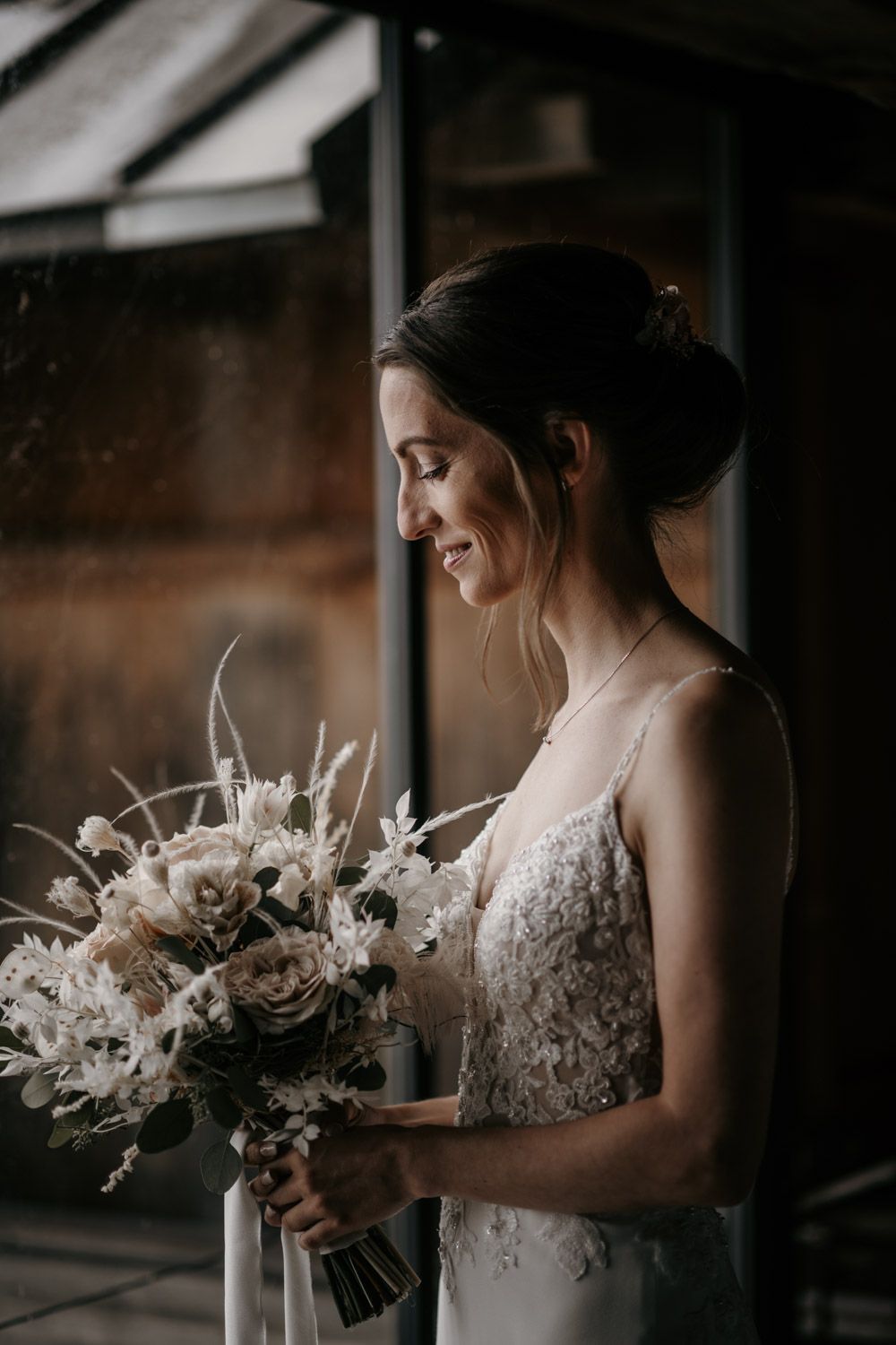A radiant bride smiles while holding a white and blush bohemian bouquet by a window.