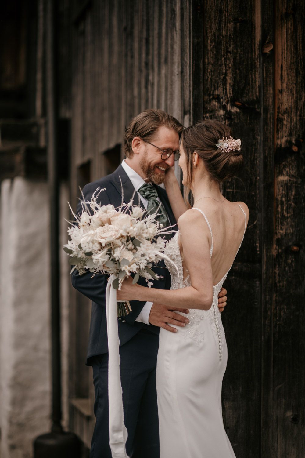 A laughing groom in a navy suit embraces his bride in a lace backless wedding gown.