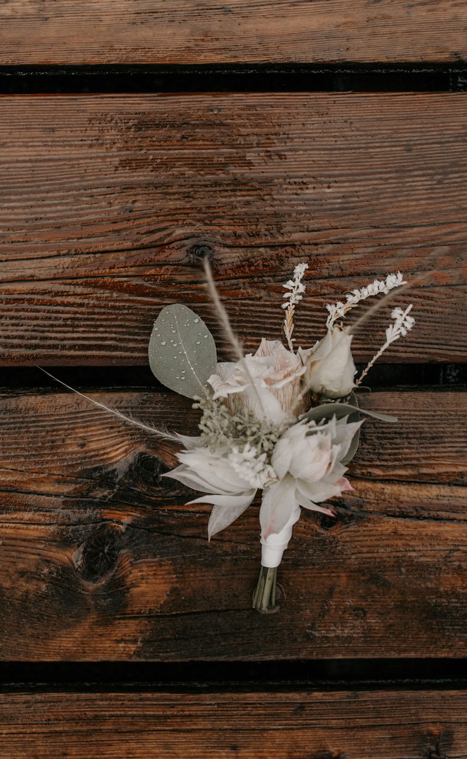 Delicate white wedding boutonniere with roses, eucalyptus, and wildflowers on rustic wood surface.