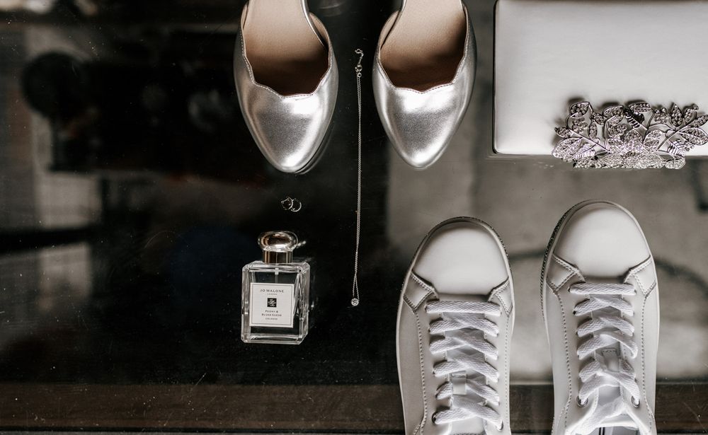 Bridal accessories flat lay featuring silver heels, white sneakers, perfume, and crystal clutch.