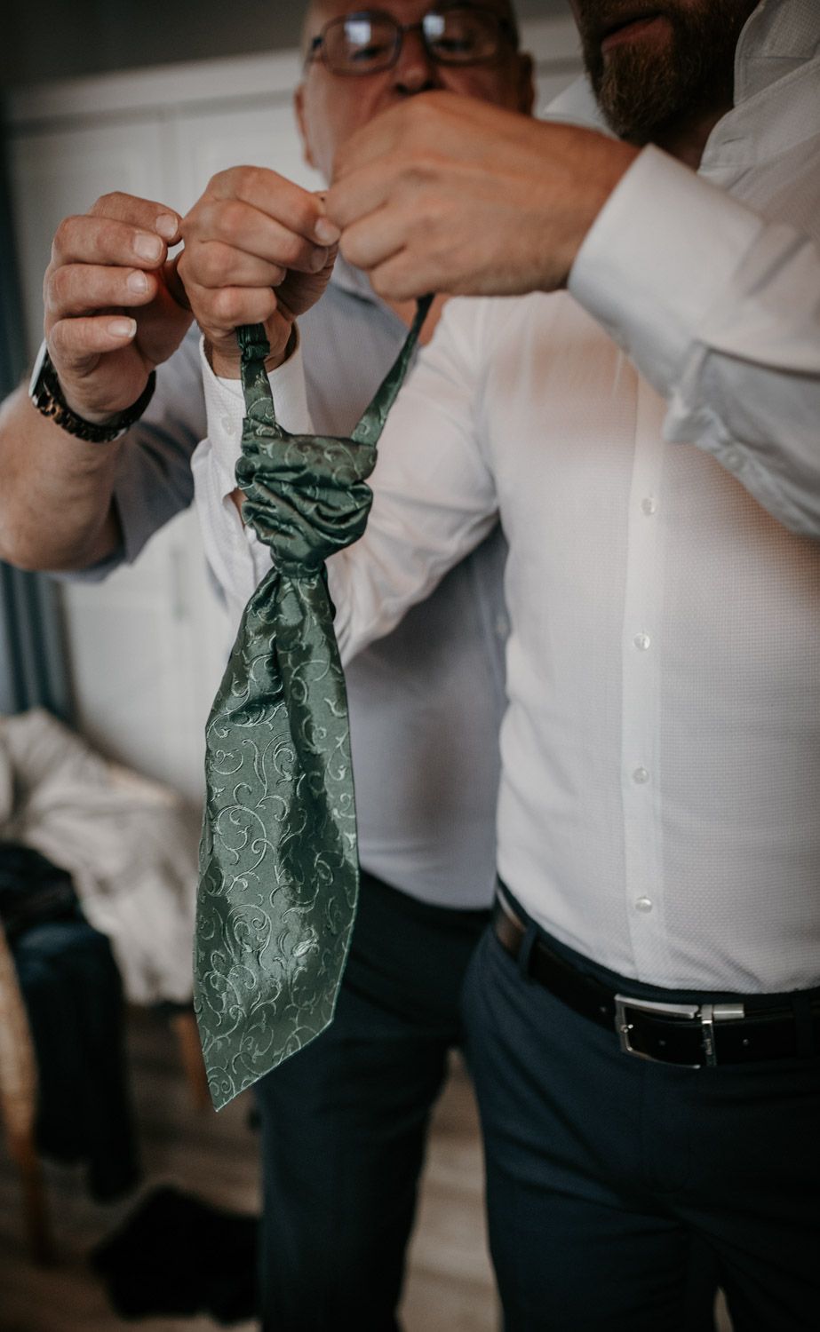 Close-up of hands tying an elegant green paisley cravat tie for a wedding groom.