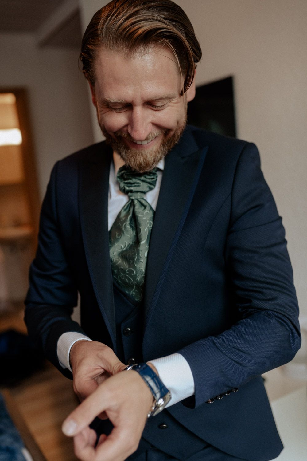 Smiling groom in navy suit adjusting cufflinks while wearing a green paisley cravat tie.