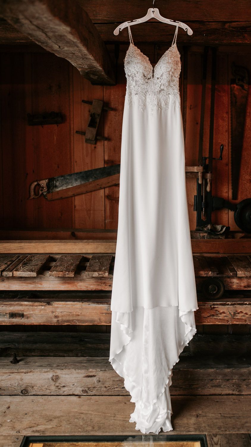 Elegant white lace bridal gown hanging in a rustic wooden barn workshop setting.