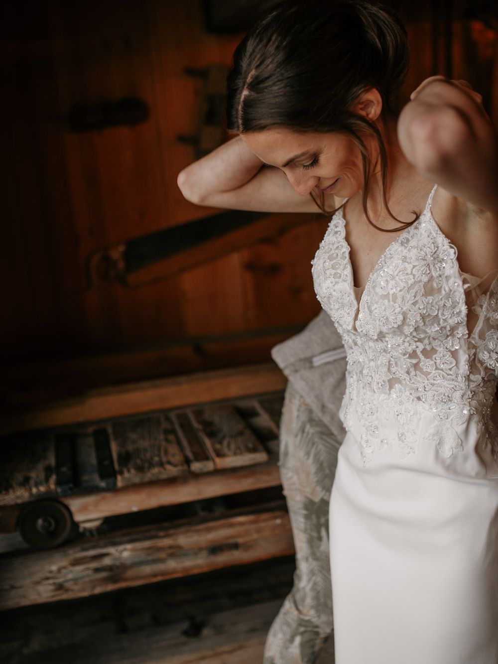 Bride in lace wedding gown adjusting her hair in a rustic wooden barn setting.