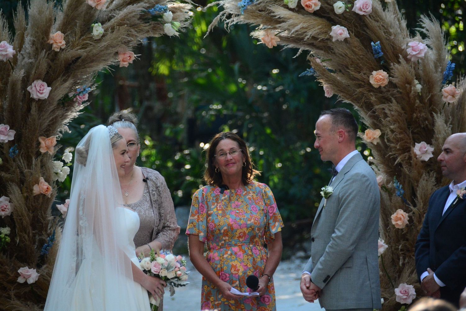 Bride and groom stand at floral pampas grass arch during outdoor garden wedding ceremony with officiant.