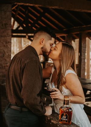 Romantic couple sharing intimate kiss while holding wine and whiskey in rustic wooden shelter.