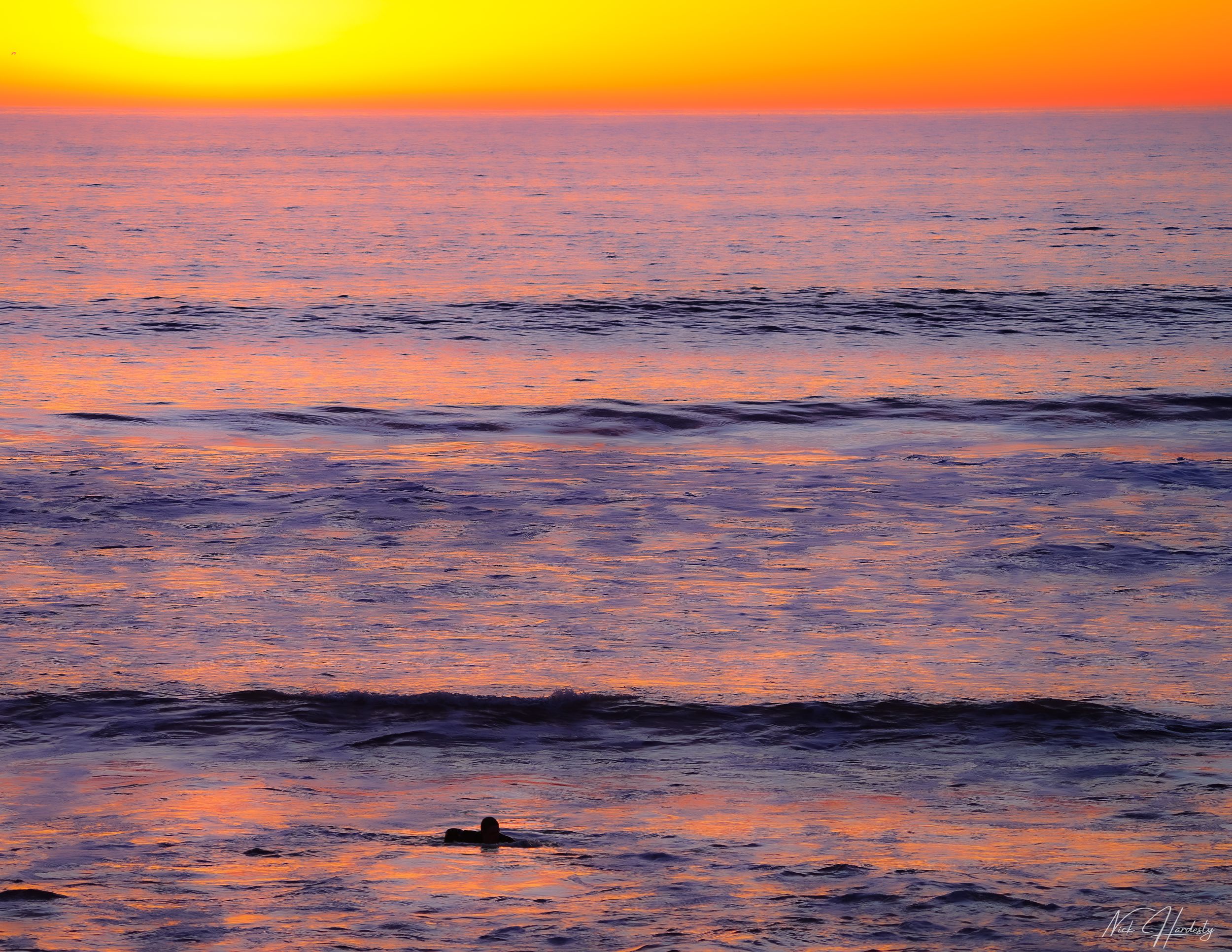 Artistic blended-color image of a surfer silhouetted against a vibrant Ventura sunset, with warm orange and purple hues merging in the sky.