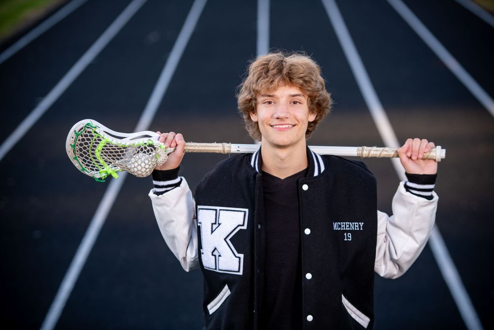 An athlete poses with lacrosse stick on running track with confident expression.
