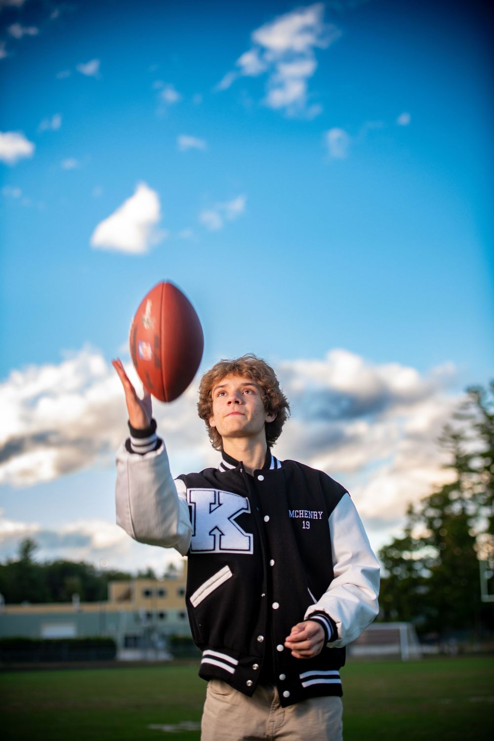 A letterman jacket-clad athlete tosses football against blue sky and clouds.