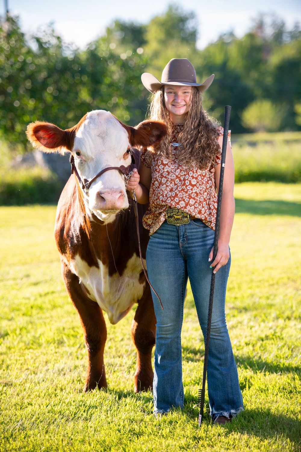 A person in cowboy hat and floral top poses with a brown and white miniature cow in a sunny field.