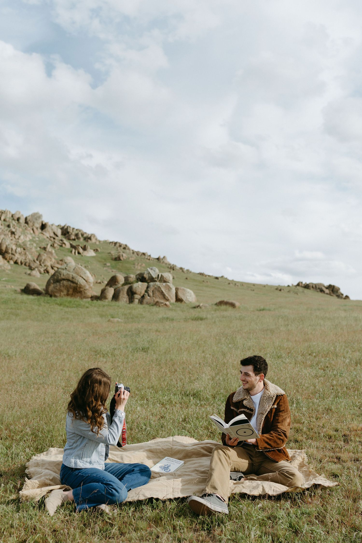 Two people having a picnic and reading on a blanket in a grassy hillside setting.