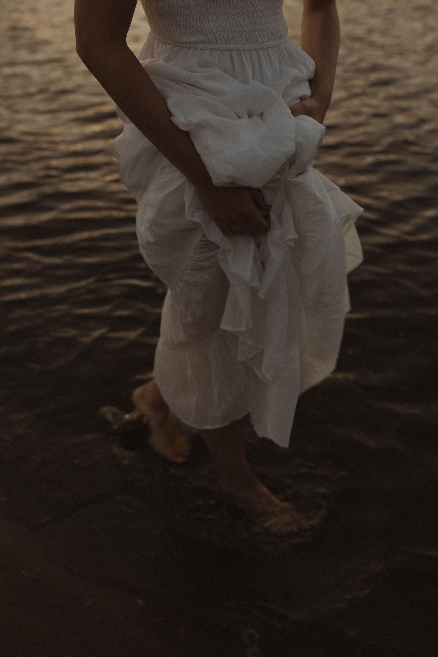 Close-up of a white wedding dress flowing in the breeze against a dark waterfront background at sunset.