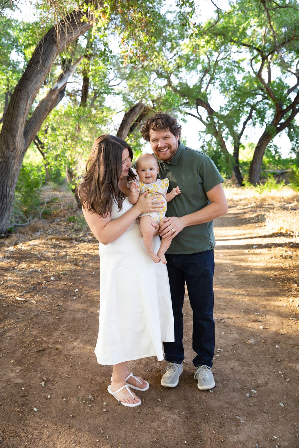 A happy young family with a baby poses on a sunny dirt path surrounded by lush green trees.