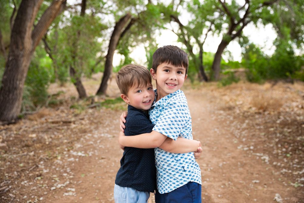 Two young boys hugging and smiling on a dirt path surrounded by green trees outdoors.