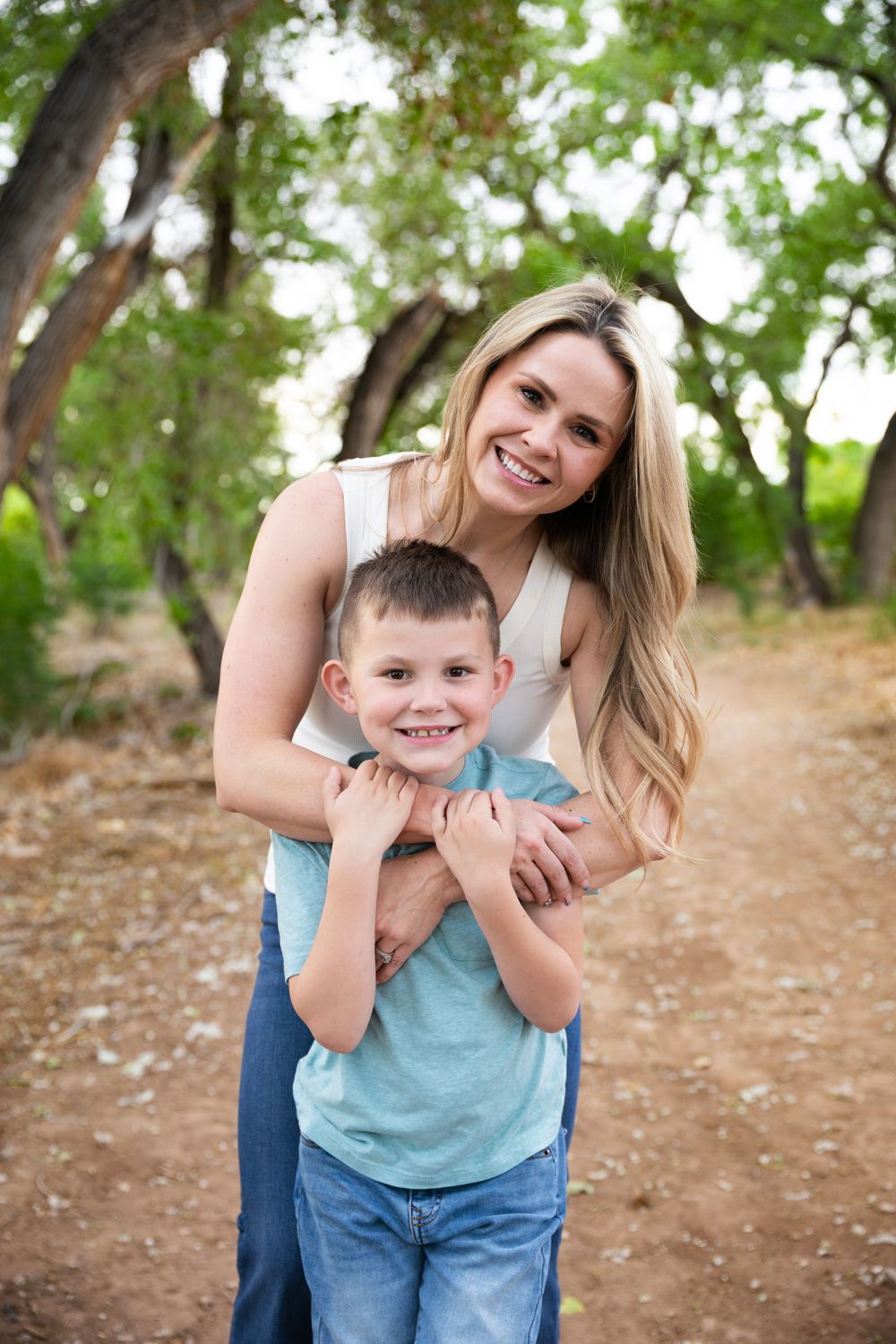 A smiling woman hugs a young boy from behind on a tree-lined dirt path outdoors.
