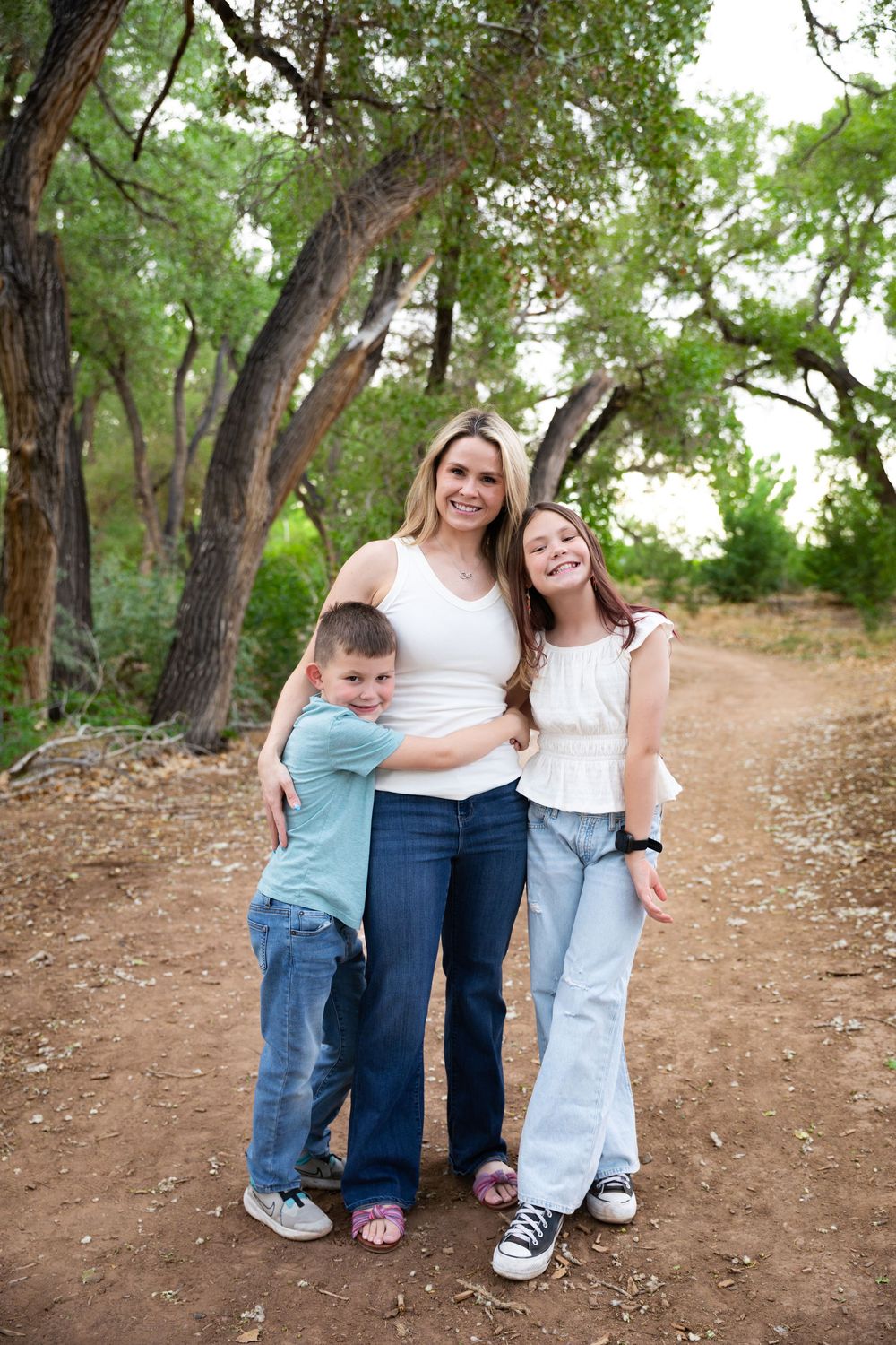 A mother smiles while her young son and daughter hug her on a dirt path surrounded by green trees.