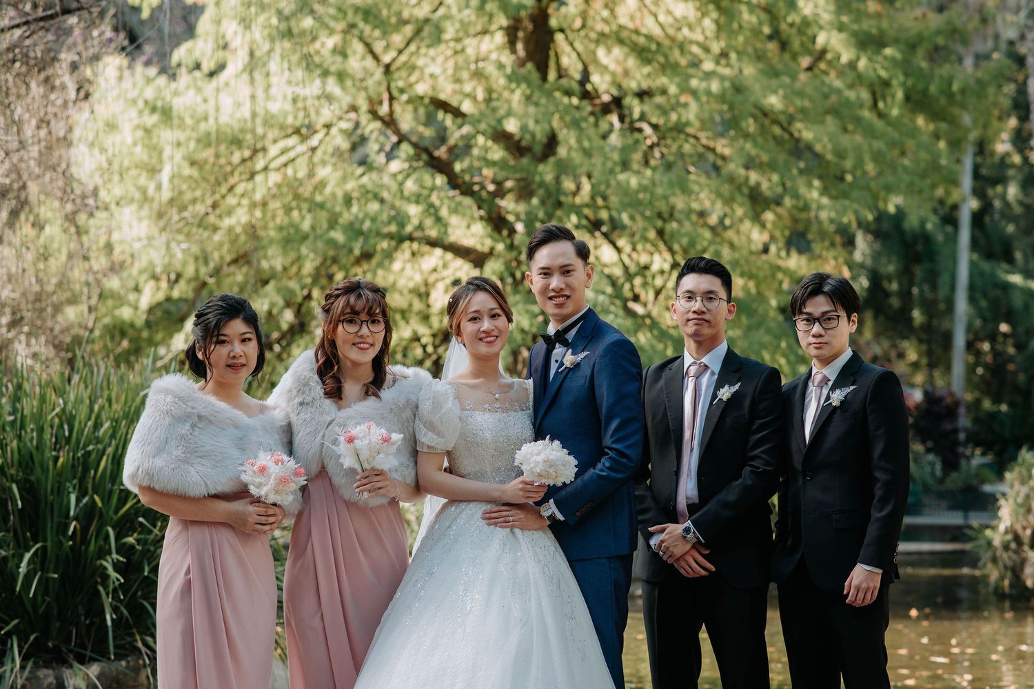 Wedding party in formal attire poses outdoors with bridesmaids in pink dresses and groomsmen in dark suits.