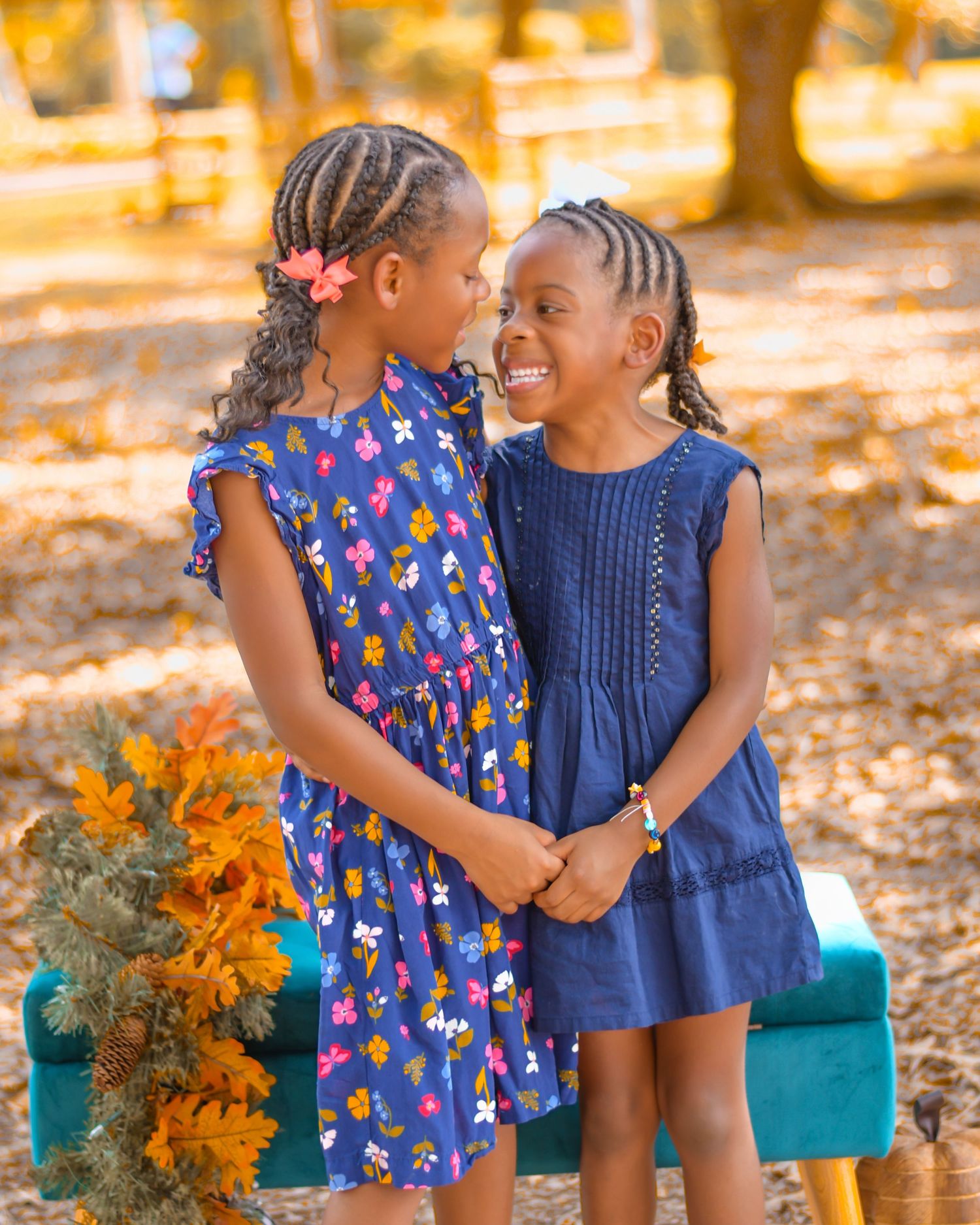 Two children in blue and floral dresses share sweet moments together among fall foliage.