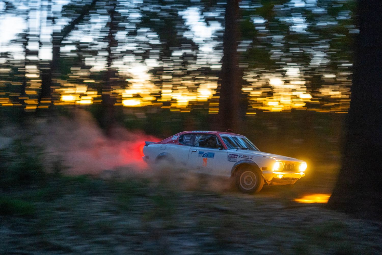 Rally car kicking up dust through forest at sunset with motion blur.