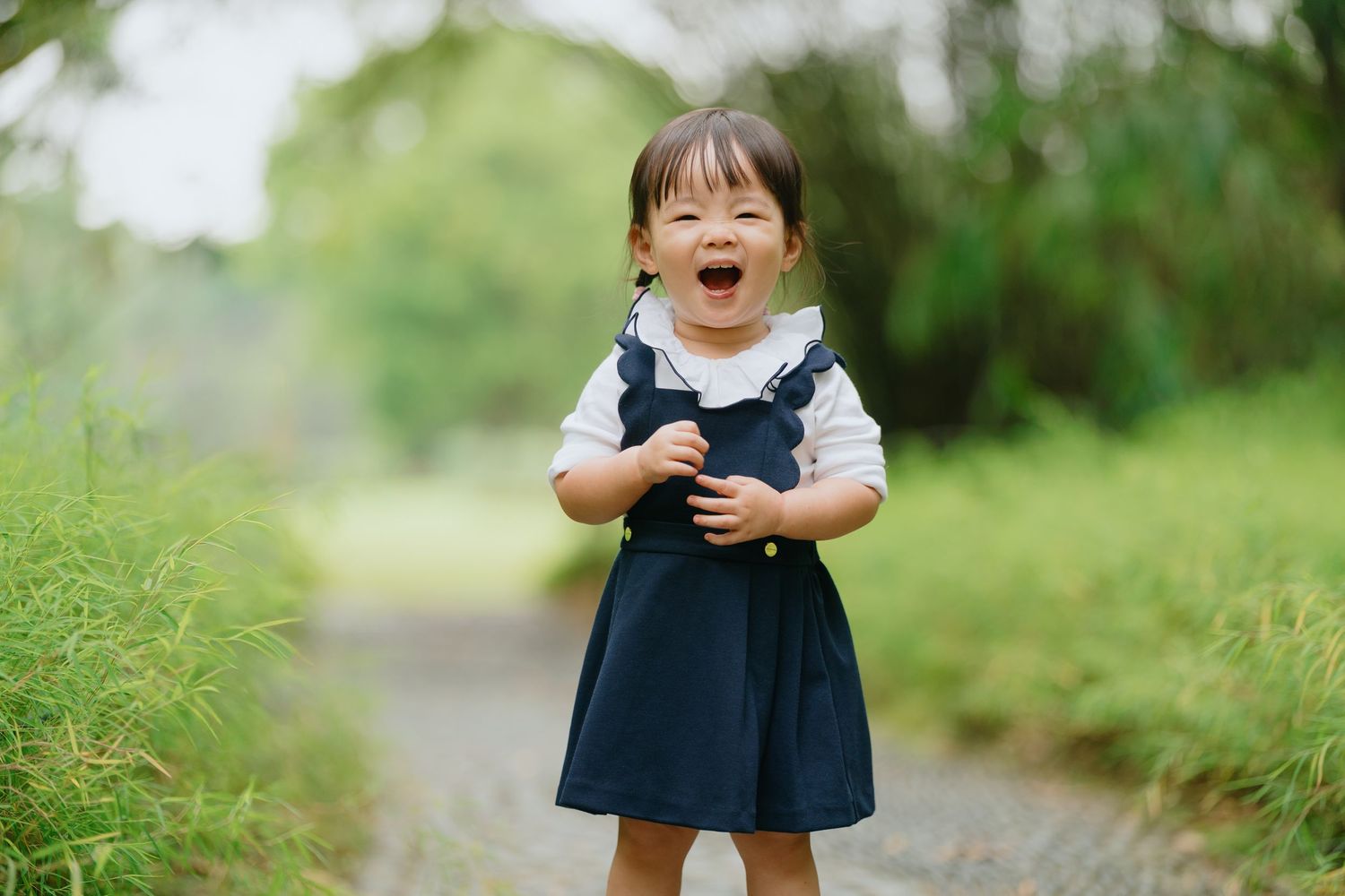 A joyful child wearing a navy blue dress plays and laughs in a lush green outdoor setting.