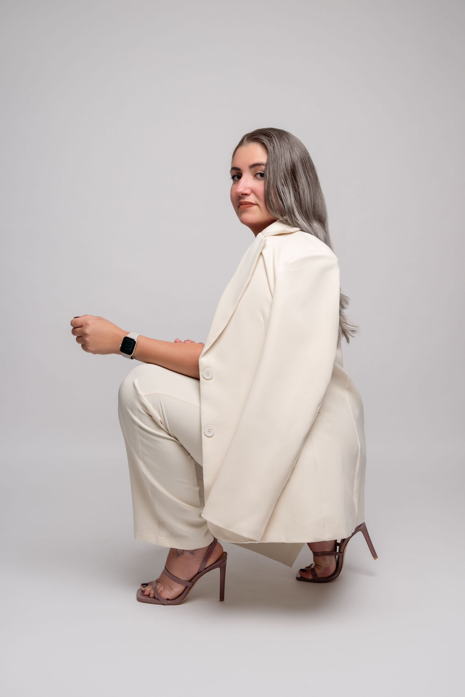 Model in cream colored business suit crouching in elegant pose against light background.