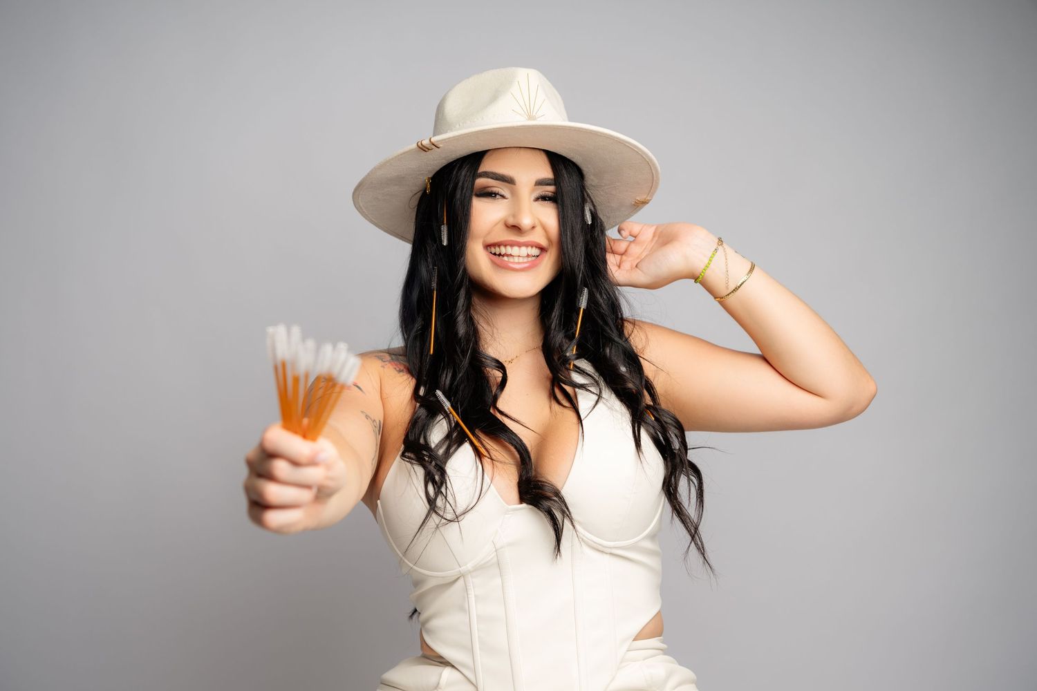 A person wearing a cowboy hat and white outfit holds a glass of alcohol while posing against a gray background.