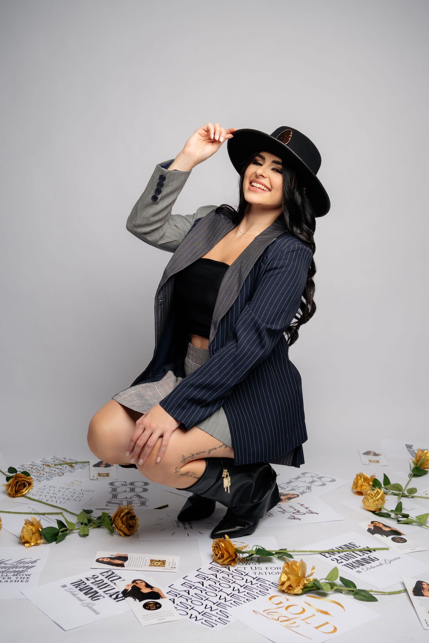 Fashion photoshoot with pinstripe blazer, black hat and boots surrounded by yellow flowers and papers on light background.