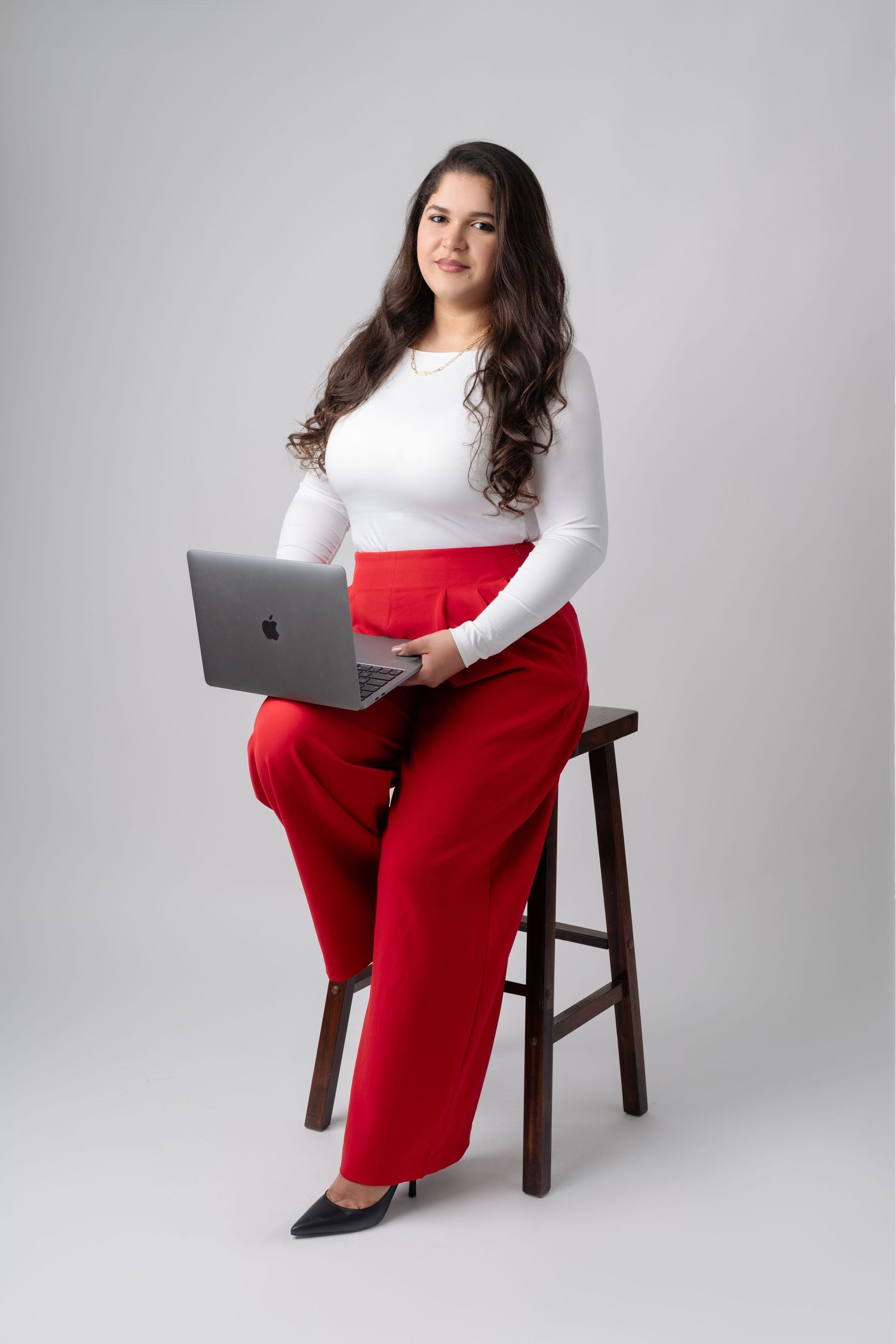 Professional woman in white top and red pants sits on wooden stool working on MacBook laptop against gray background.