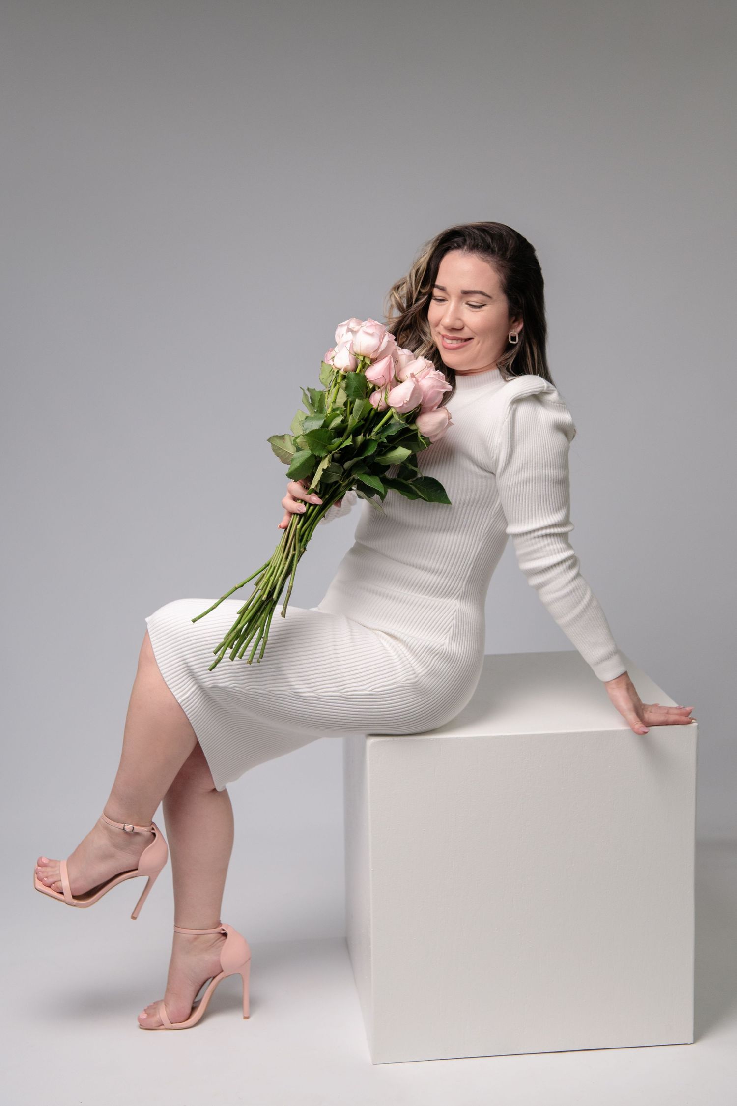 Woman in white dress holding pink roses while sitting on white cube in studio setting.