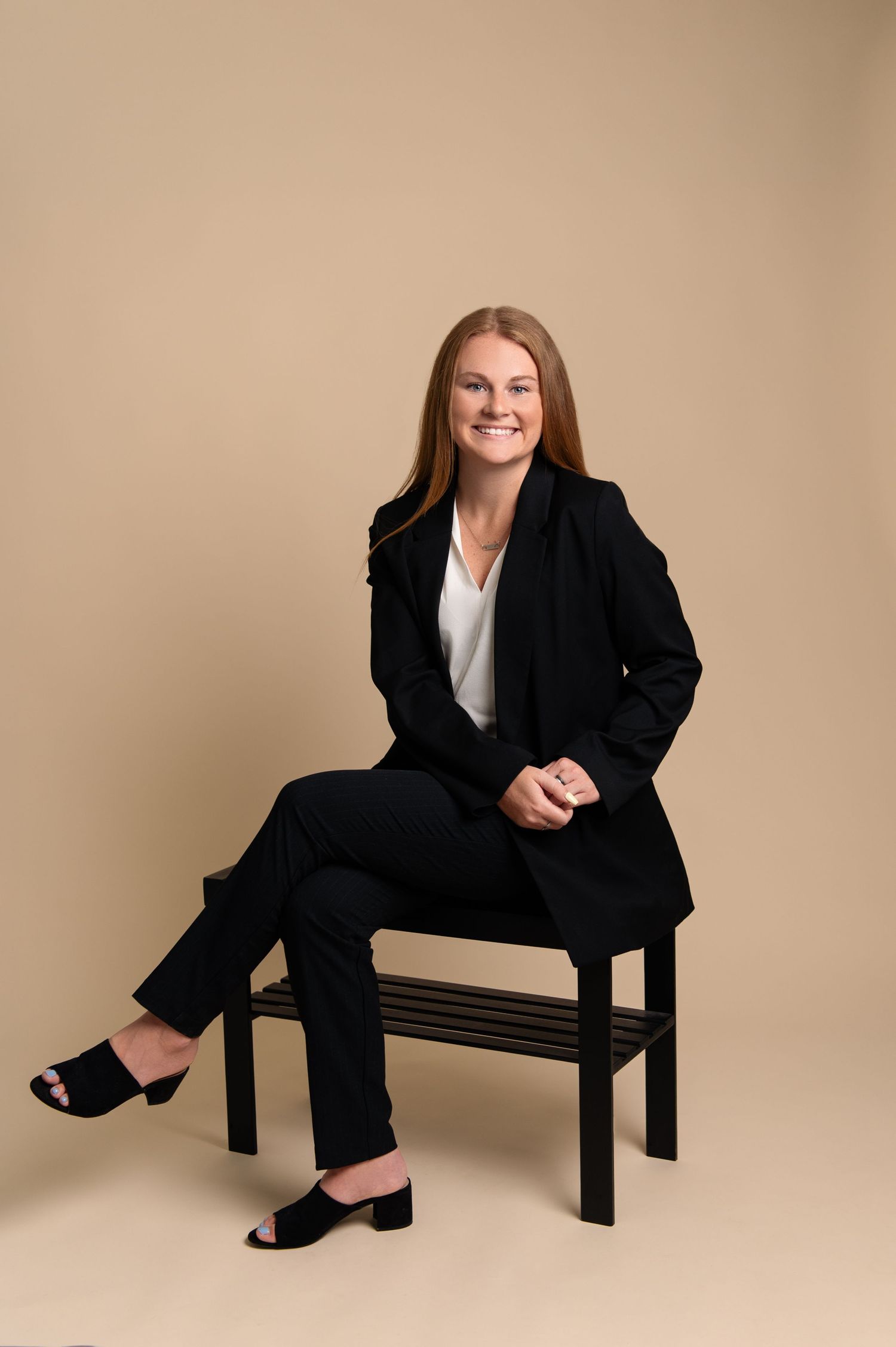 Professional portrait of someone in business attire seated on a wooden bench against beige backdrop.