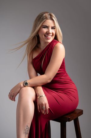 Fashion portrait of a woman in elegant red dress sitting on wooden stool against gray backdrop.
